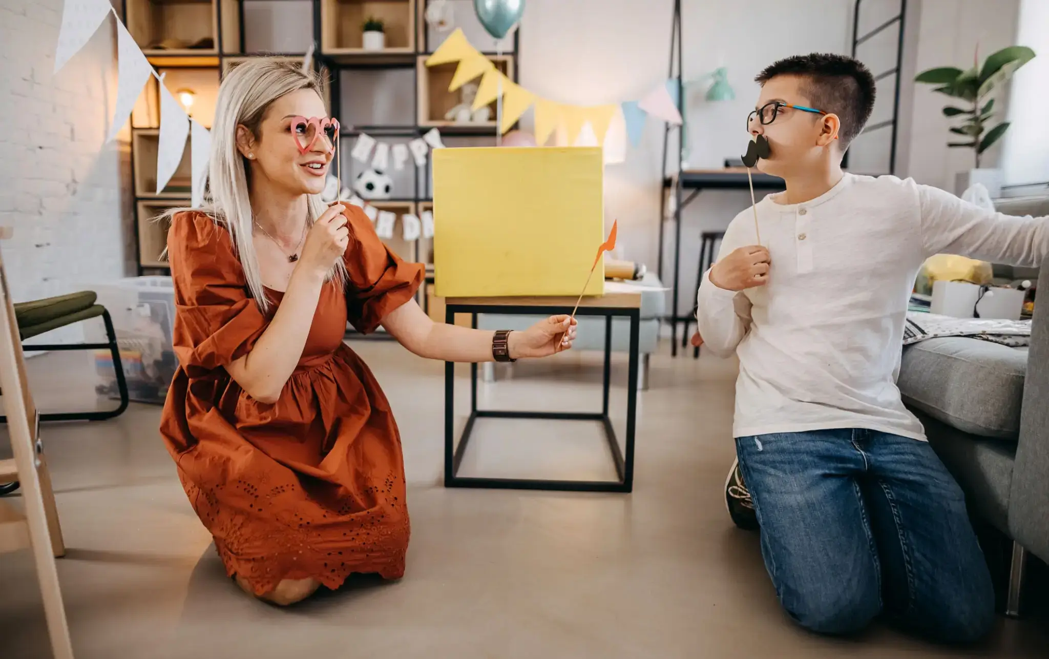 Two colleagues cheer with coffee mugs near a yellow box in a cozy office.