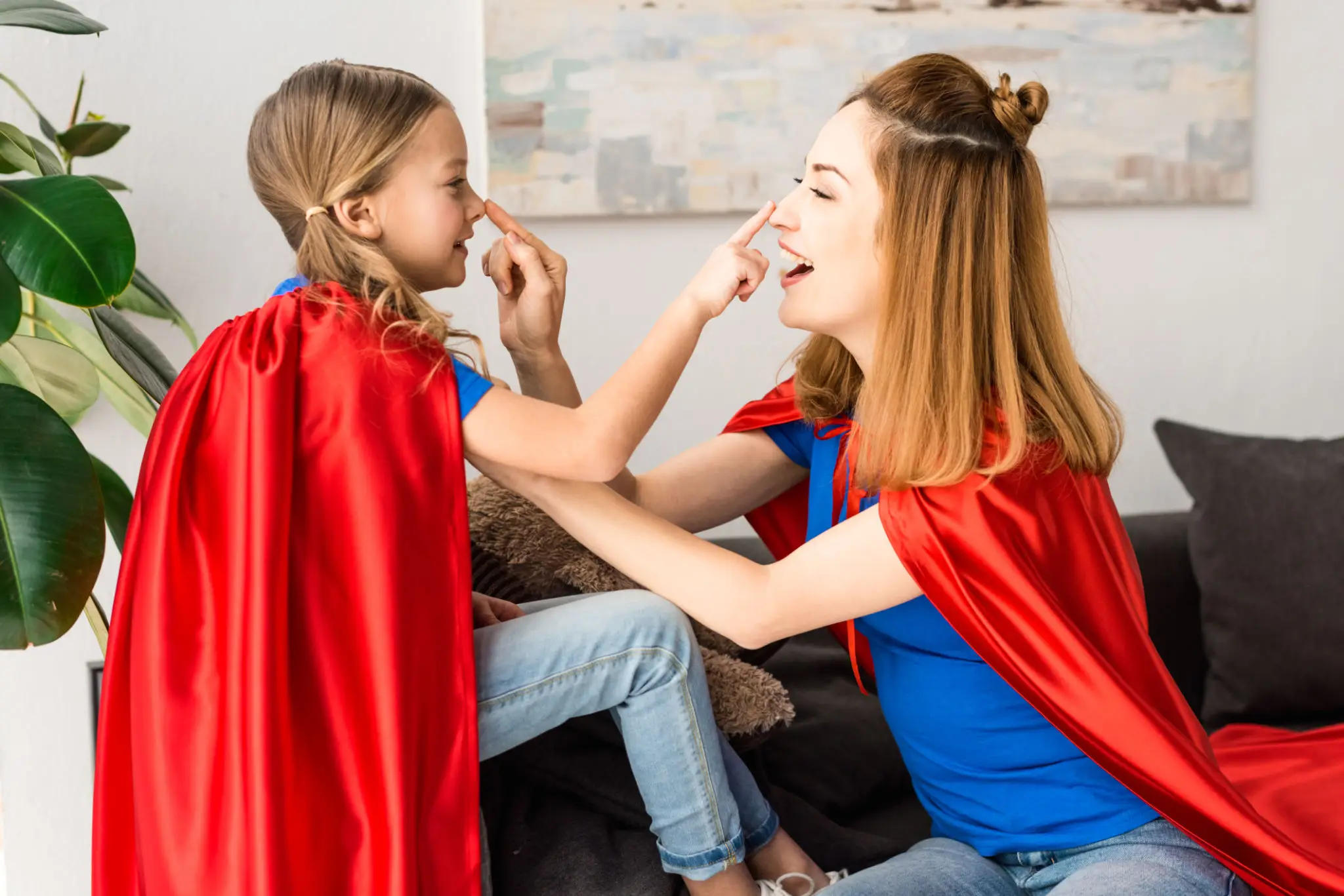 Woman and girl in red capes playfully touching noses indoors.